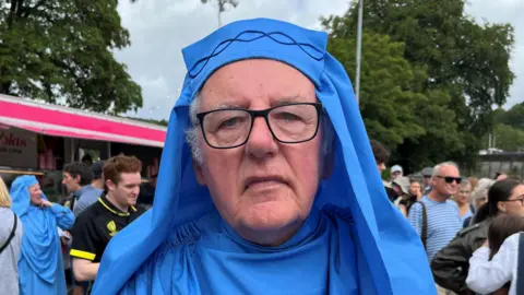 BBC An older man wearing light blue druid robes and dark glasses. 