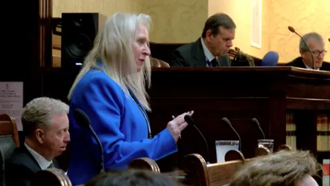 Onchan MHK Julie Edge has long blonde straight hair. We see her side-on facing the right, standing in the Tynwald chamber, where members sit around her. She is wearing a blue suit jacket.