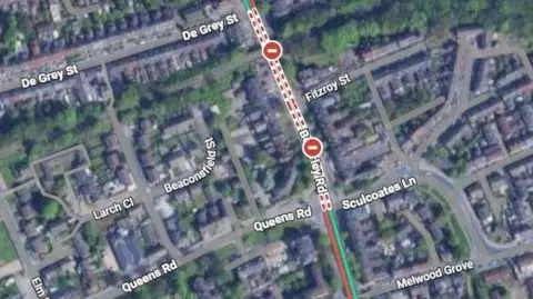 Google A Google Maps picture of a street which has red lines going down it to signify it is closed and an incident is happening. Text can be seen around it for surrounding streets which provide an aerial view of the area.