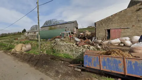 A crumbling farm on the side of a road. There are hay bails and trailers and a falling down dry stone wall.