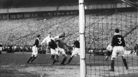 A black‑and‑white action shot shows multiple players contesting the ball in front of goal during a crowded match. The goalpost and net dominate the right side, with a defender wearing number three positioned near the line. Behind the scene, a packed Hampden Stadium forms a dense backdrop and a sea of people.
