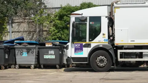 A bin lorry next to some skips which are full.