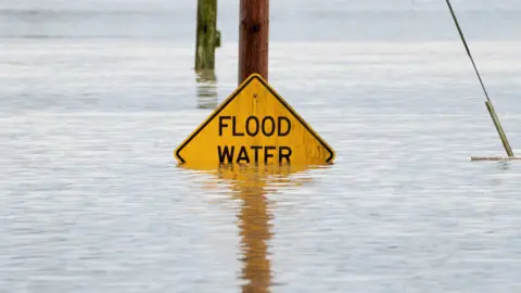 A stock image of a flood water sign partly submerged by water. It is a yellow diamond shaped sign attached to a wooden pole. Just under half the sign is under water.