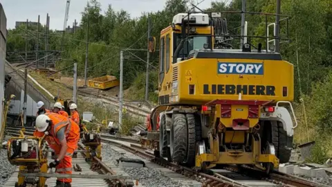 A large yellow construction vechicle is stationary on the right hand side of the tram track. A construction worker wearing an orange high-vis jacket and a white safety helment is using a piece of yellow construction machinary. Three other construction workers also wearing orange high-vis jackets and white safety helmets are standing in the distance.