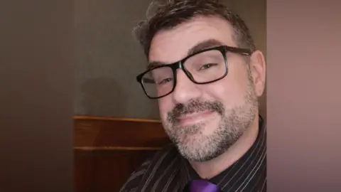 Handout A man with greying hair and beard with glasses. He is wearing a striped suit and purple tie.