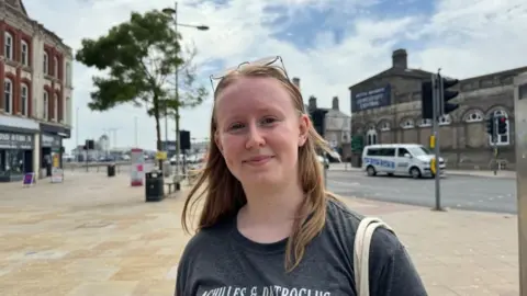 Andrew Turner/BBC Katie Plant has long blonde hair, and her sunglasses are rested on her head. She is wearing a black tee shirt, and has bag straps over her left shoulder. She is pictured on the Station Square in Lowestoft, with Lowestoft Central railway station in the backdrop with the former Tuttles Department Store on the left of the image.