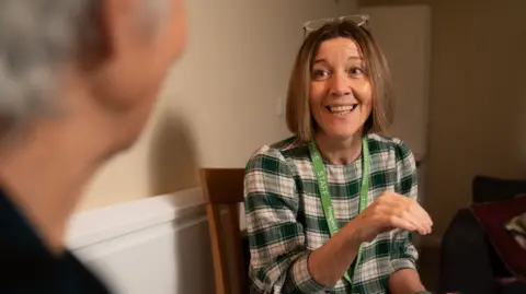 Prospect Hospice A woman talking to another woman while smiling. The woman who is talking has brown hair in a long bob haircut and she is wearing a green and white checked top. She has glasses balanced on her head and she is wearing a green lanyard that says 'staff' on it. The other woman is to the left of the picture and is out of focus.