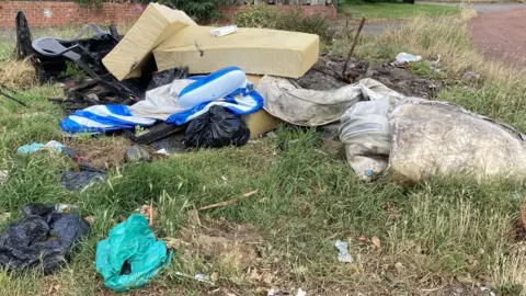 A heap of rubbish including the remains of a mattress, foam blocks, a broken plastic chair and a number of bags of waste on a grassed area with what appears to be the remains of a fire.
