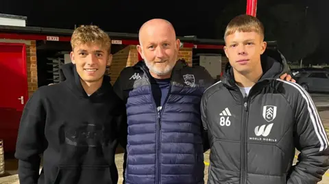 Paul Renton Callum, Paul, and Luca are standing side by side at night outside a sports venue, wearing casual and athletic jackets, with kegs and a red building entrance in the background.