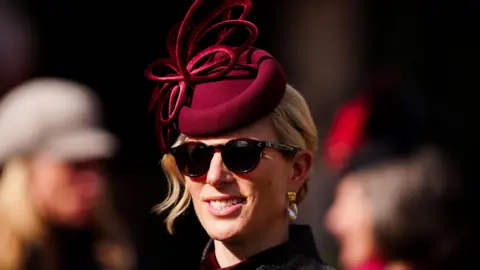 PA Media Zara Tindall wears sunglasses and a red felt hat with a red fascinator on top as she arrives at Cheltenham 2026