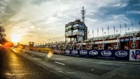STEVE BABB The sun rises over a stretch of road next to a red brick grandstand with long rows of benches. There is a taller building with windows which stands in the centre and banners have been draped around it. Race banners can also be seen lining barriers next to the tarmac.