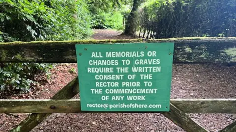 A weathered wooden gate at the entrance to a graveyard supports a green sign with white lettering stating that all memorials or changes to graves require the written consent of the rector, with a narrow path and dense greenery visible beyond.