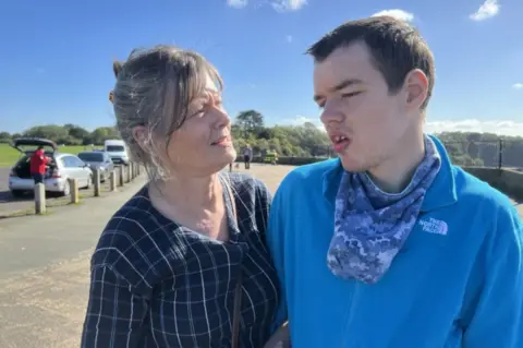 A woman and young man stand close to one another on a sunny day. The woman has grey hair held up by a tortoiseshell clip and with some hair around her face. She is wearing a navy and white checked blouse and has a handbag over her shoulder. She is looking into the eyes of a young man who is looking off camera. He has short brown hair and is wearing a light blue North Face fleece and blue patterned scarf.