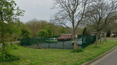 A car park next to a residential road with trees and grass all around.