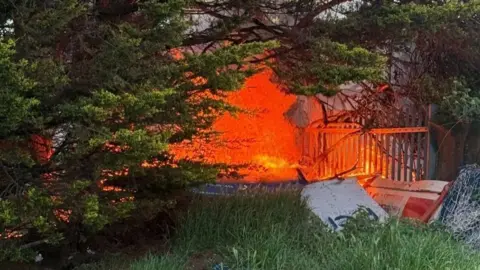 Sunderland RCA A small orange blaze in a wooden area by a fence. The blaze is underneath some trees. 