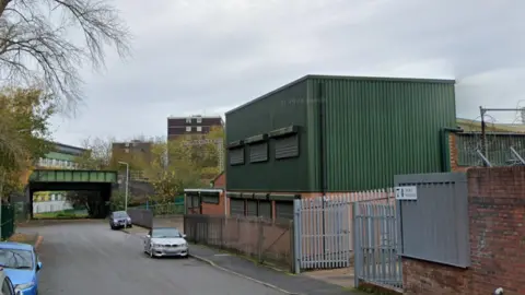 Google Maps A street with green and redbrick warehouse buildings on the right side, behind a metal fence. A railway bridge can be seen towards the end of the street.