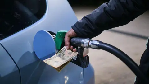 A hand holding a petrol pump into a car as it fills up at a petrol station in Zimbawe. The same hand also holds paper money. The car is silver.