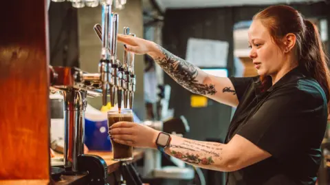 A female bartender pouring a pint of draught beer from the tap behind the bar in a busy British pub.