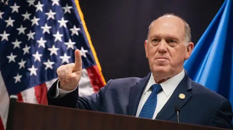 Tom Homan, White House border czar, stands in front of an American flag wearing a blue suit, white shirt and blue tie