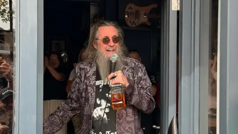 A man with long grey hair and beard. he has round tinted glasses on and is holding a microphone and a bottle of whiskey in his hand. 