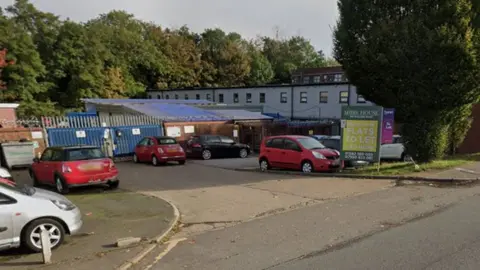 The entrance to a set of buildings on an industrial estate. Three red cars are parked sporadically and there are trees to the side and in the background. One building has small windows running all along. There are blue metal gates set back from the road.