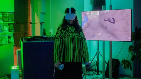 Dreamy Place A person with long curly black hair wearing a green and black vertical stripy shirt. The person is holding two white controllers in their hands and is wearing a white VR headset on their head. They are stood in front of a TV screen in a green room