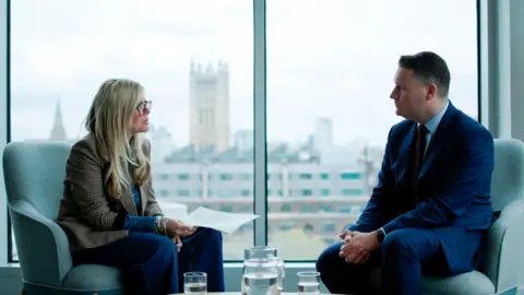 The BBC's Emma Barnett sits on the left in an armchair holding some papers while to the right is former Health Secretary Wes Streeting sitting opposite. He has short brown hair and wears a navy suit, a blue shirt and a dark tie. He clasps his hands together, while behind them Parliament's Victoria Tower can be seen through the window, rising up over the rooftops.