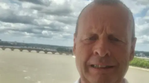 David Munster Selfie taken by David of him standing against a railing above a brown murky river which stretches on behind him with a bridge in the distance. He smiles at the camera. 