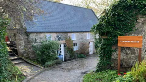 A granite brick building with a granite cobbled road and a sign which says La Moinerie Village