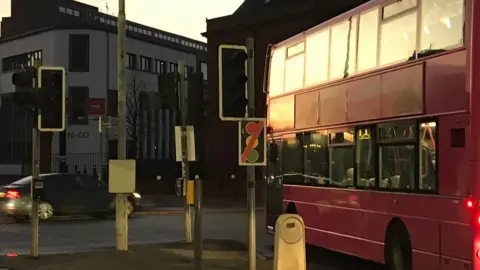 Traffic lights at Ormeau Road/Cromac Street/Ormeau Avenue out of action on Friday morning.  A pink Metro bus is waiting at the malfunctioning lights while a car passes through the junction.  There is a warning sign at the junction which shows a traffic light signal with a red diagonal line through the picture, to indicate the lights are not working. 