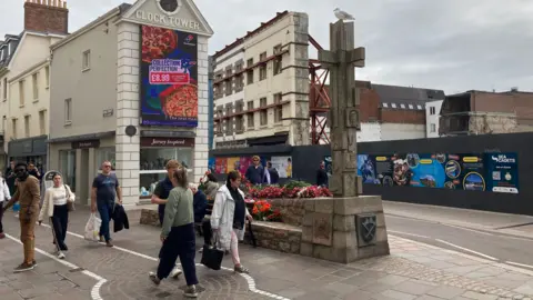 BBC People walking past Charing Cross in St Helier, Jersey. A cross statue is on a plinth on the street. A building site with hoarding around its edge is on the opposite side of the road from the cross. A large video screen on a building called the Clock Tower has an advert for Domino's pizza on it.