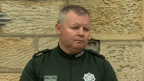 Supt Robert McGowan speaking to reporters at a press conference, He has short, grey hair and is wearing a green police t-shirt.