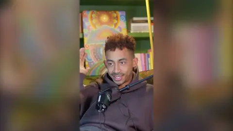 Jordan has short curly hair and is wearing a dark purple hoodie.