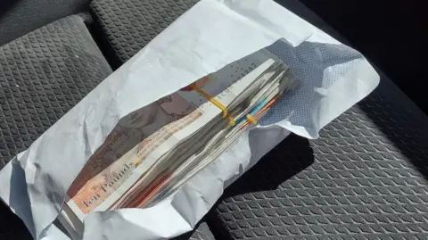 Oxfordshire County Council A white envelope with several bands of banknotes on a car seat.