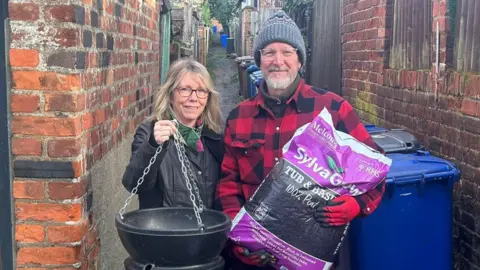 two people stand in an alley with a bag of compost and a hanging basket.