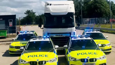 Four marked police cars parked alongside a white HGV lorry cab at an industrial estate. The HGV cab towers above the cars, making it easier for police officers to see into the cabs of other lorries. 