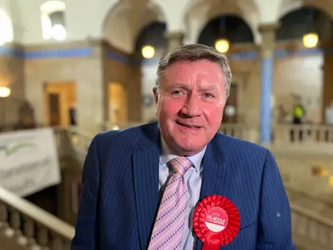 Dennis Jones looking at the camera. He is wearing a blue stripy suit, a pink patterned tie and a large red Labour rosette. In the background is the interior of a council building.