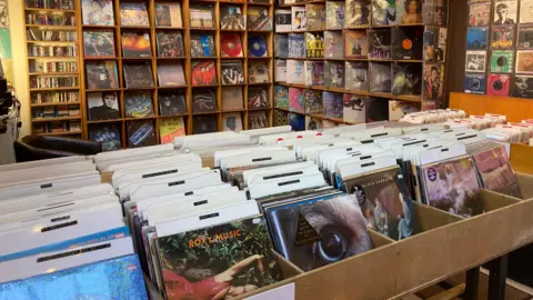 A large number of vinyl records in their sleeves are on shelves and in drawers in a record shop.