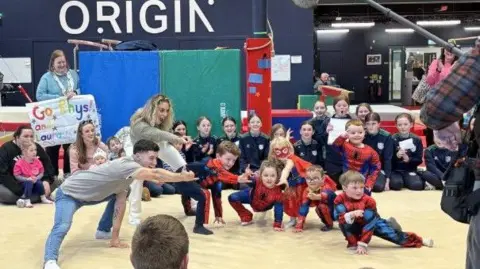 A group of people posing of the camera with a group of six children who are dressed in red Spiderman costumes