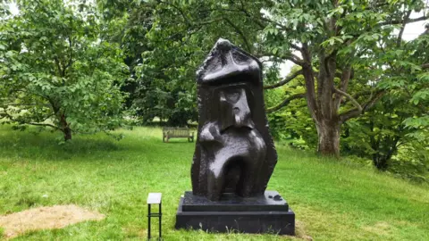A semi-abstract bronze sculpture on display at a botanic garden. There are trees in the background. 