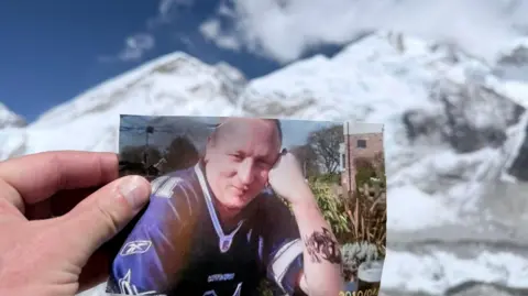 Eugene Scardifield A photograph of a smiling man in a blue sports shirt is held by a hand in front of snow covered mountains