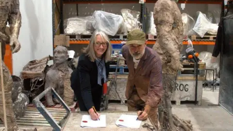East Suffolk Council Caroline Topping and Laurence Edwards pictured in his foundry. They lean over a table, each with a white paper contract in front of them, and they both have a pen in their hand. They look at the camera and smile. Caroline has long grey hair and wears a black top and trousers. Laurence wears a green hat and a purple shirt.