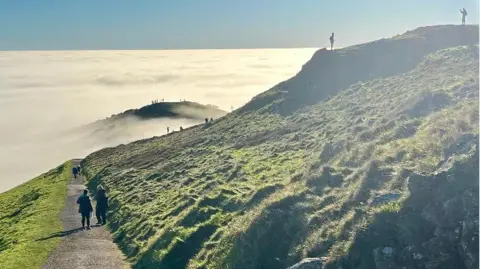 BBC Weather Watchers/Toppo People are walking on a footpath amid the hills as well as on the top of them. Cloud surrounds the hills below but blue skies can be seen.