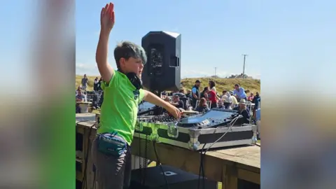 Family Photo Efan stands behind a DJ deck in front of a crowd. Green hills and a blue sky can be seen in the background. He wears a bright lime green shirt and has blue in his hair. He has one hand up in the sky and another on the deck.