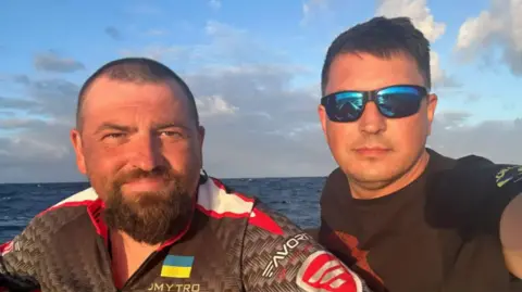 Dmytro Rezvoy Dmytro Rezvoy, in blue sunglasses and a black T-shirt, and Ivan Havrylko, wearing a shirt with Ukraine's flag on it, in their rowing boat at sea. 