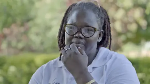 Evelyn Amon wearing glasses and a white shirt holds her hand up towards her mouth