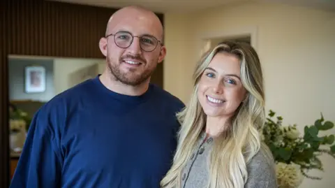 A man and woman stand close together both smiling into camera. The man has a shaved head, is wearing glasses and a dark blue t-shirt. The woman has long blonde hair, blue eyes and is wearing a grey cardigan.