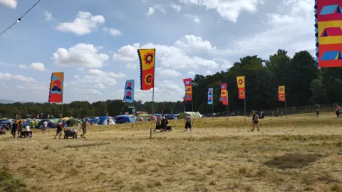 A camping field at Kendal Calling. People are pulling trolleys full of their belongings along a field with browned grass. There are colourful flags dotted around the site and camping tents in the background.