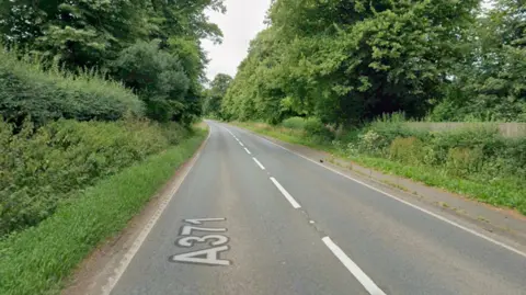 An A-road is pictured with one lane either side. Both sides have grass verges, with bushes and trees above.