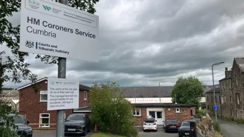 Cartel a la entrada del tribunal forense de Cockermouth. Es un letrero blanco pintado sobre un poste de metal plateado, que presenta varios logotipos y nombres de agencias gubernamentales, con la letra más destacada en el centro que dice Oficina forense de Cambria. El hastial del edificio de ladrillo de dos pisos con techo de tejas puntiagudo se encuentra en ángulo recto con una adición de un piso con techo plano que parece albergar la entrada principal. Hay una tienda de Sainsbury's detrás de un pequeño edificio. Los vehículos estarán estacionados afuera de la oficina del forense. Al otro lado de la calle estrecha y sin pavimentar, a la derecha del edificio, hay edificios de piedra mucho más antiguos de dos y tres pisos en terrazas.